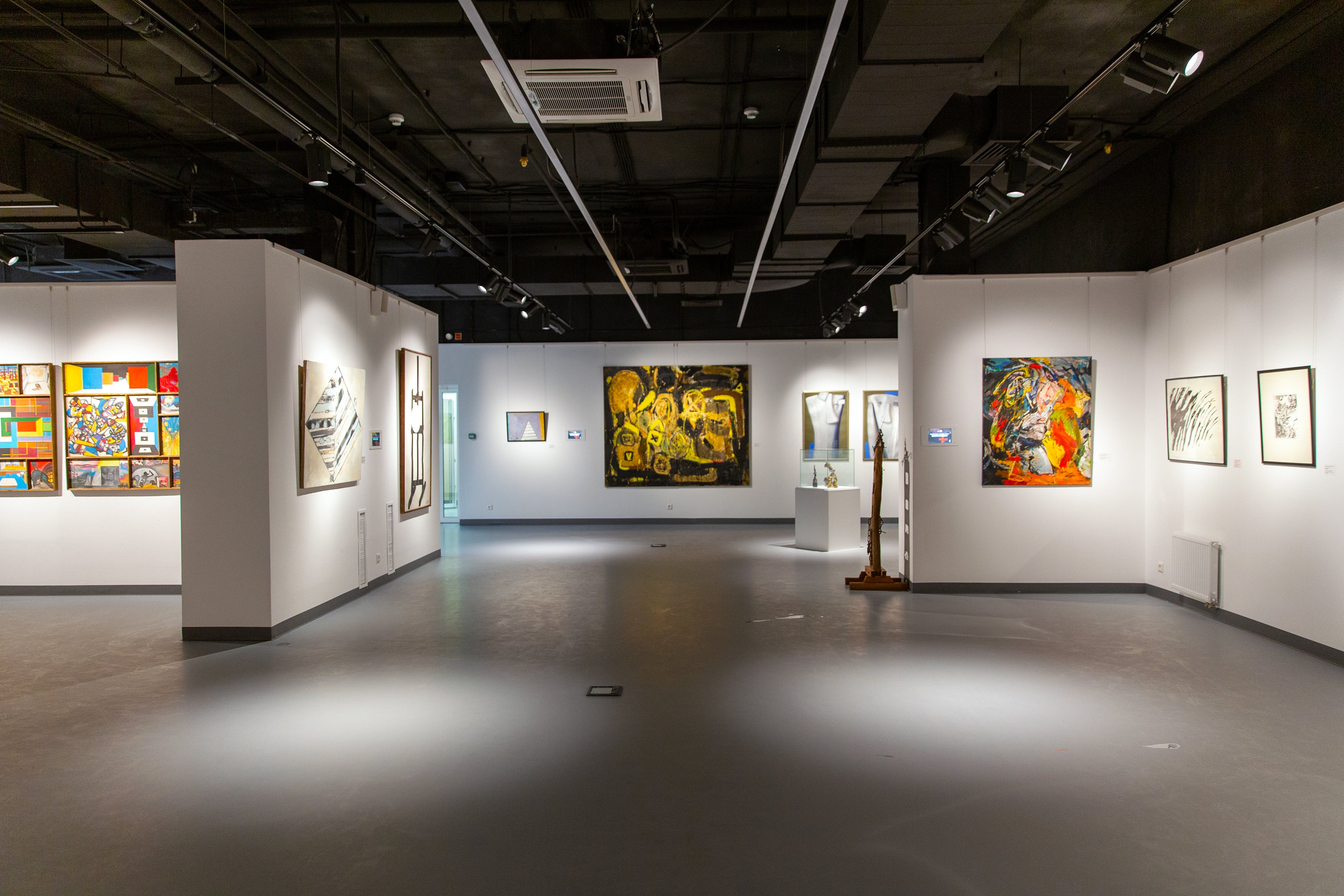 Spacious contemporary art gallery interior showcasing a diverse collection of artwork, with brightly lit walls featuring a mix of colorful abstract paintings, intricate sculptures, and monochrome prints. The focal point is a large golden abstract canvas in the center, flanked by a variety of expressive pieces, all thoughtfully curated to provide visual engagement and cultural enrichment for visitors.